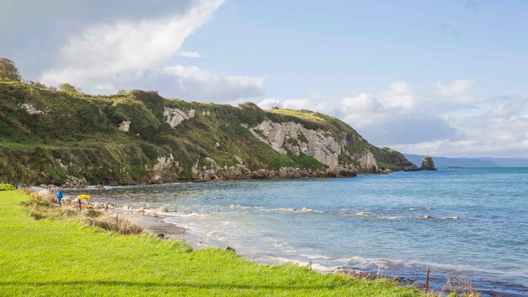 A view towards grass-covered cliffs along the Portmuck coastline, Islandmagee peninsula, County Antrim. Two visitors are visible walking along the shoreline in the distance.